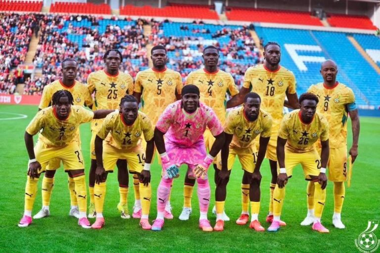Soccer team in yellow kits posing for a group photo on the field, with a pink goalkeeper standing center front.