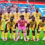 Soccer team posing for a photo on the field; all players in yellow uniforms, goalkeeper in pink kneels center front with teammates standing behind him in a stadium.
