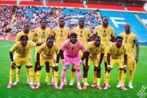 Soccer team posing for a photo on the field; all players in yellow uniforms, goalkeeper in pink kneels center front with teammates standing behind him in a stadium.