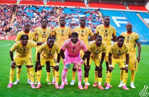 Soccer team posing for a photo on the field; all players in yellow uniforms, goalkeeper in pink kneels center front with teammates standing behind him in a stadium.