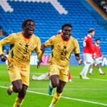 Two Ghanaian soccer players in yellow uniforms celebrate running across a bright green pitch as fans watch from blue stands in the background.