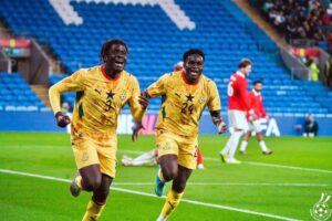 Two Ghanaian soccer players in yellow uniforms celebrate running across a bright green pitch as fans watch from blue stands in the background.