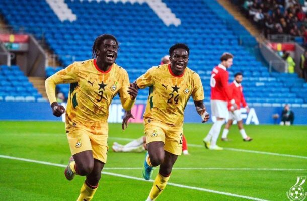 Two Ghanaian soccer players in yellow uniforms celebrate running across a bright green pitch as fans watch from blue stands in the background.