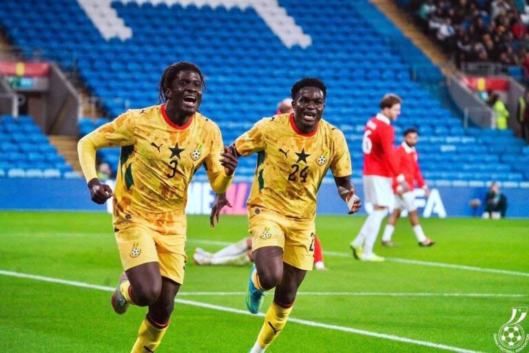 Two Ghanaian soccer players in yellow uniforms celebrate running across a bright green pitch as fans watch from blue stands in the background.
