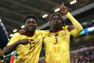 Two soccer players in yellow uniforms celebrate on the field, arms around each other after a goal, with a stadium crowd in the background.