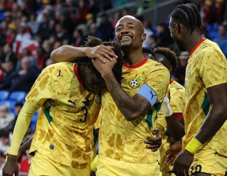 Football players in yellow jerseys celebrate on the field, hugging and smiling after a goal surrounded by teammates in a crowd.
