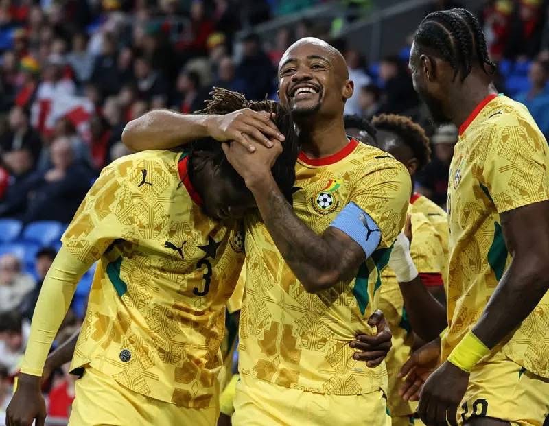 Football players in yellow jerseys celebrate on the field, hugging and smiling after a goal surrounded by teammates in a crowd.