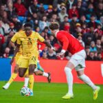 Soccer players compete for the ball on a green field as a crowd watches from the stands, one in yellow and one in red.