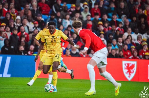 Soccer players compete for the ball on a green field as a crowd watches from the stands, one in yellow and one in red.