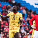 Two footballers on a field, one in a yellow Ghana kit (number 18) gesturing, the other in a red Portugal kit looking on. Crowd in stands behind them.