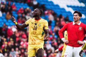 Two footballers on a field, one in a yellow Ghana kit (number 18) gesturing, the other in a red Portugal kit looking on. Crowd in stands behind them.