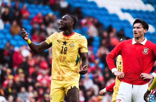 Two footballers on a field, one in a yellow Ghana kit (number 18) gesturing, the other in a red Portugal kit looking on. Crowd in stands behind them.