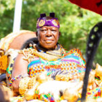Man in vibrant traditional attire seated outdoors, wearing a purple headband and ornate jewelry, participating in a cultural ceremony.
