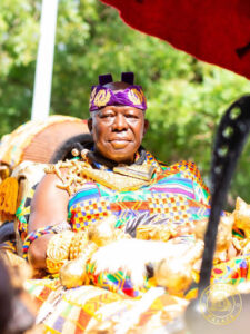 Man in vibrant traditional attire seated outdoors, wearing a purple headband and ornate jewelry, participating in a cultural ceremony.