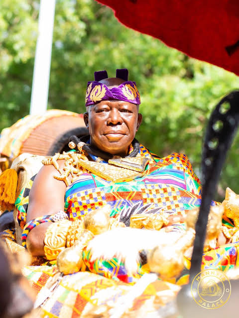 Man in vibrant traditional attire seated outdoors, wearing a purple headband and ornate jewelry, participating in a cultural ceremony.