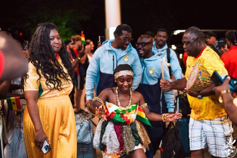 Young girl in colorful traditional dress dances while surrounded by cheering people and travelers, flags in hand at a festive event at night (Ghanaian colors).