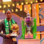 Man in dark traditional outfit and green scarf posing with a gold football-shaped trophy, beside a large silver trophy with green ribbons on a stage.