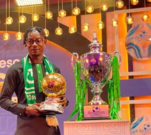 Man in dark traditional outfit and green scarf posing with a gold football-shaped trophy, beside a large silver trophy with green ribbons on a stage.