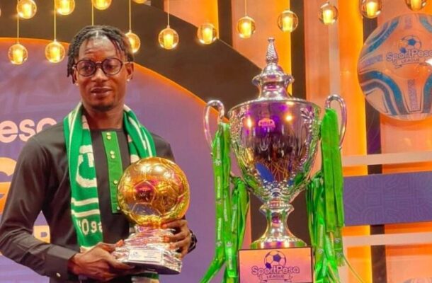 Man in dark traditional outfit and green scarf posing with a gold football-shaped trophy, beside a large silver trophy with green ribbons on a stage.