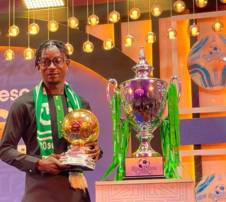 Man in dark traditional outfit and green scarf posing with a gold football-shaped trophy, beside a large silver trophy with green ribbons on a stage.
