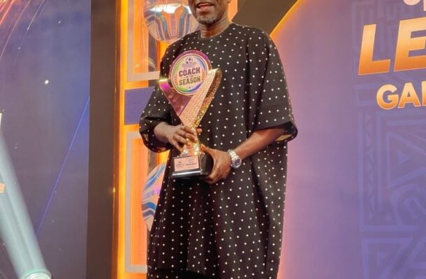 Man in traditional agbada holding a 'Coach of the Season' trophy on stage with warm hanging lights in the background with a purple/orange backdrop.