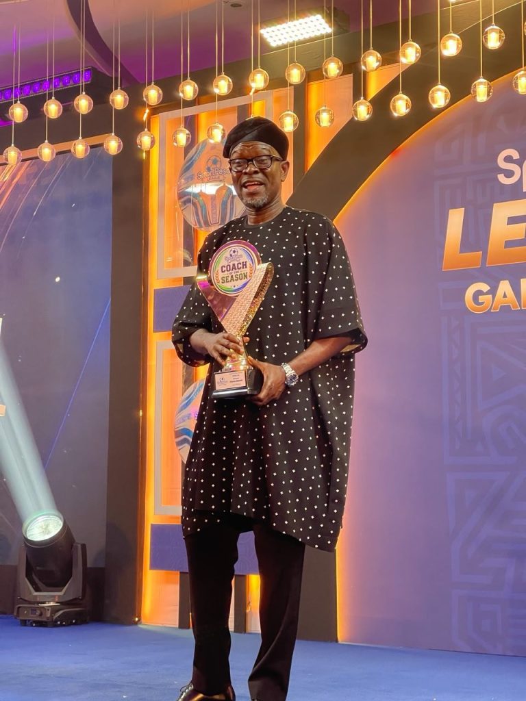 Man in traditional agbada holding a 'Coach of the Season' trophy on stage with warm hanging lights in the background with a purple/orange backdrop.