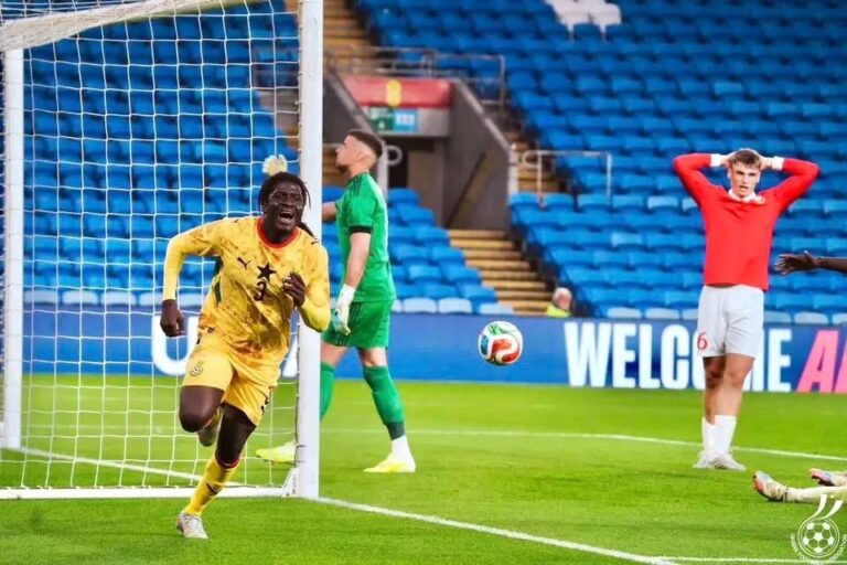 Soccer player in yellow jersey runs past the goal as a white net and empty blue stands appear, with a green-clad goalkeeper and a distressed red-shirted player in the background, suggesting a goal-scoring moment.