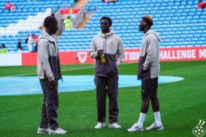Three young men in matching gray hoodies stand on a football pitch, one holding a water bottle as they chat.