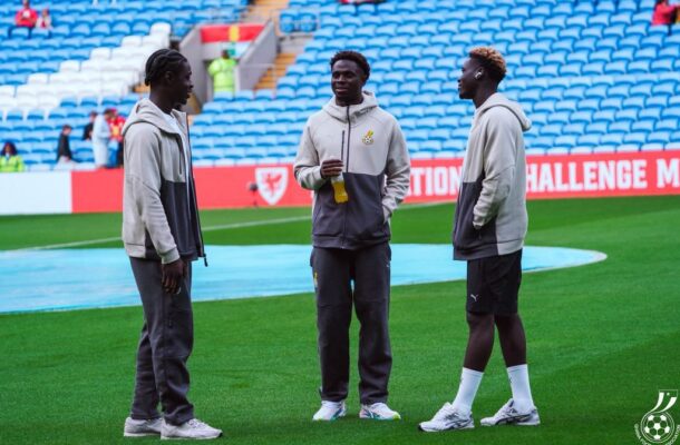 Three young men in matching gray hoodies stand on a football pitch, one holding a water bottle as they chat.