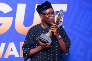 Man in a dark patterned shirt and glasses kisses a tall trophy against a blue backdrop.