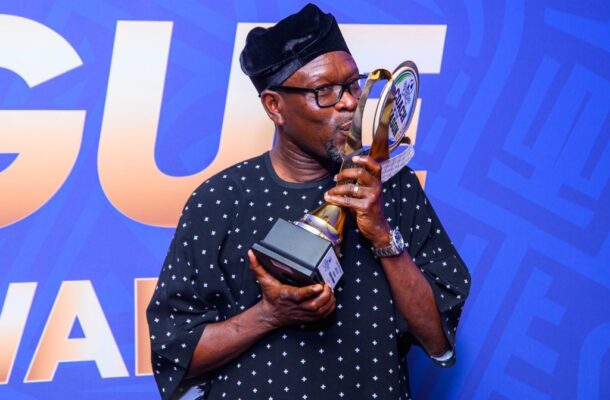 Man in a dark patterned shirt and glasses kisses a tall trophy against a blue backdrop.