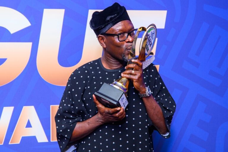 Man in a dark patterned shirt and glasses kisses a tall trophy against a blue backdrop.