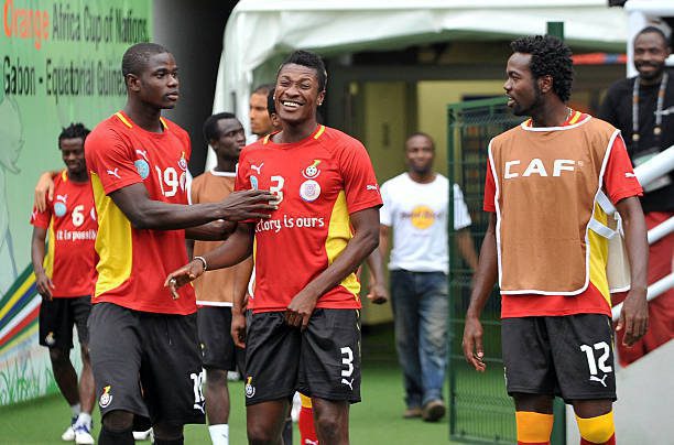 Ghanaian football players in red training kits celebrate together near a tunnel/dugout area on the field.