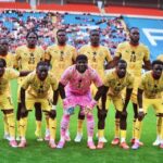 Soccer team in yellow uniforms poses for a pre-match photo on a green field with stands full of fans in the background; goalkeeper wearing pink in front center.