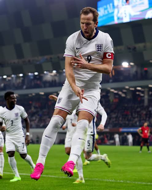 England captain wearing white kit with armband jumps in celebration on a football pitch with pink boots during a match