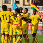 Soccer players in yellow jerseys celebrate a goal on the field, with numbers 11, 20, 23 visible; flags in the stands.