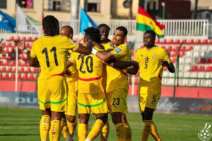 Soccer players in yellow jerseys celebrate a goal on the field, with numbers 11, 20, 23 visible; flags in the stands.