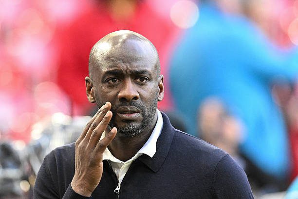 Man with a shaved head and stubble, wearing a dark polo, touching his face with a worried expression; blurred crowd in the background.