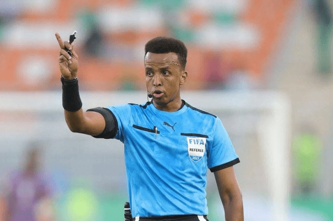 Referee in a blue kit raises a whistle and points during a soccer match, signaling a call.