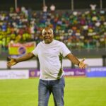 Man in a white shirt on a soccer field with arms outstretched, cheering crowds in the background.