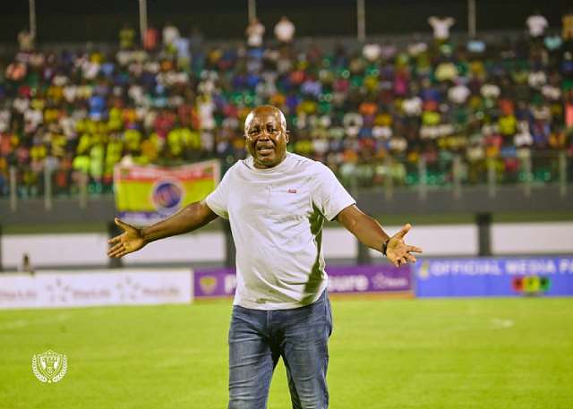 Man in a white shirt on a soccer field with arms outstretched, cheering crowds in the background.