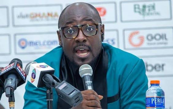 IMG_0875 | Ghanaguardian.com Man in a teal jacket speaks into a microphone at a press conference, with sponsor logos on the backdrop and multiple microphones in front.