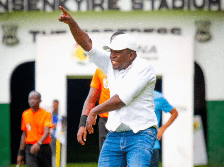 Man in a white polo and cap points forward while celebrating on a football pitch near the tunnel entry behind him