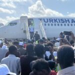 Crowd of people with phones raised greeting passengers as a Turkish Airlines plane arrives and people exit via portable stairs.