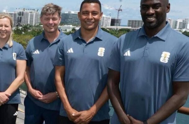 Four adults in matching blue polo shirts pose for a team photo on a balcony, with a city skyline and water in the background.