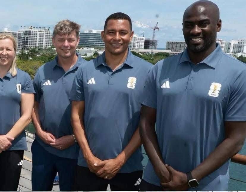 Four adults in matching blue polo shirts pose for a team photo on a balcony, with a city skyline and water in the background.