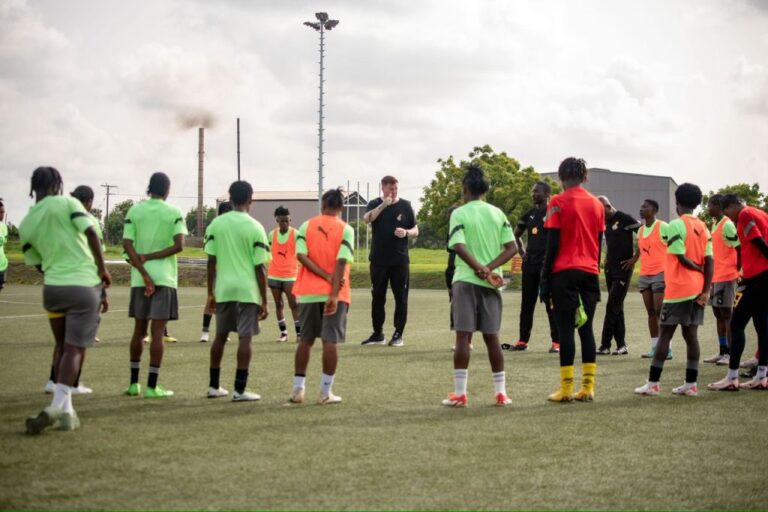 Soccer players stand in a circle on a grassy field as a coach speaks during practice, wearing neon green and orange bibs.