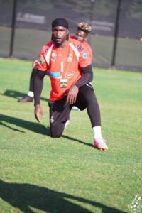 Soccer goalkeeper in an orange jersey with number 1 kneeling on a grassy field during training; black sleeves and headband, with another player in the background.