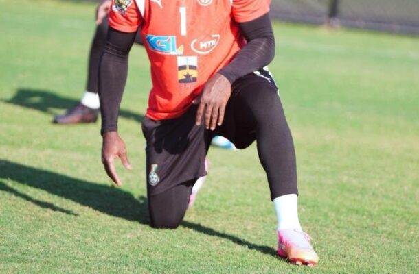Soccer goalkeeper in an orange jersey with number 1 kneeling on a grassy field during training; black sleeves and headband, with another player in the background.