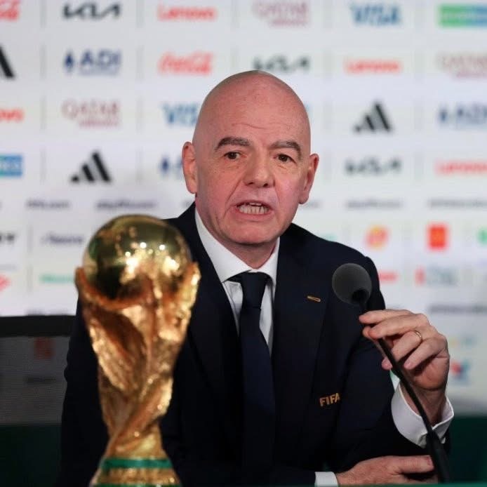 Bald man in a dark suit speaks into a microphone at a press conference, with a golden FIFA World Cup trophy in the foreground.
